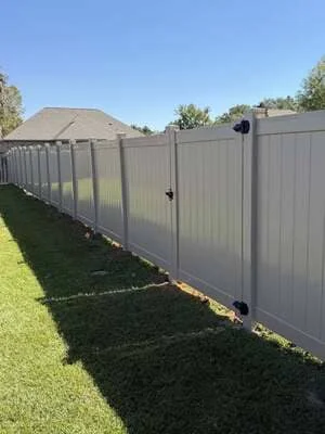 Gray vinyl privacy fence installed along residential property with green lawn and trees in background